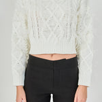 Mid-shot view of cream long-sleeve cable-knit sweater focusing on the crew neckline, wide ribbed cropped hem, and feather detailing.