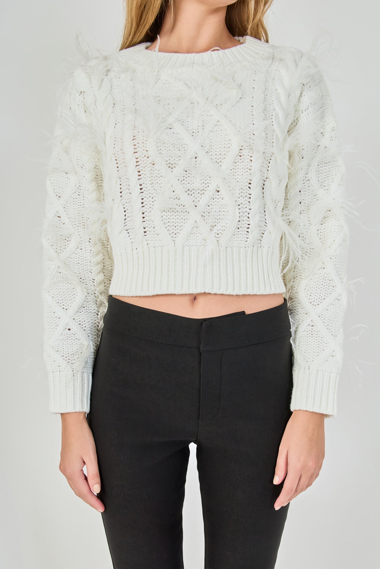 Mid-shot view of cream long-sleeve cable-knit sweater focusing on the crew neckline, wide ribbed cropped hem, and feather detailing.