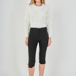 Full-length front view of a cream cropped cable-knit sweater with feather accents, styled with black fitted capri trousers.