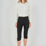 Full-length front view of a cream cropped cable-knit sweater with feather accents, styled with black fitted capri trousers.