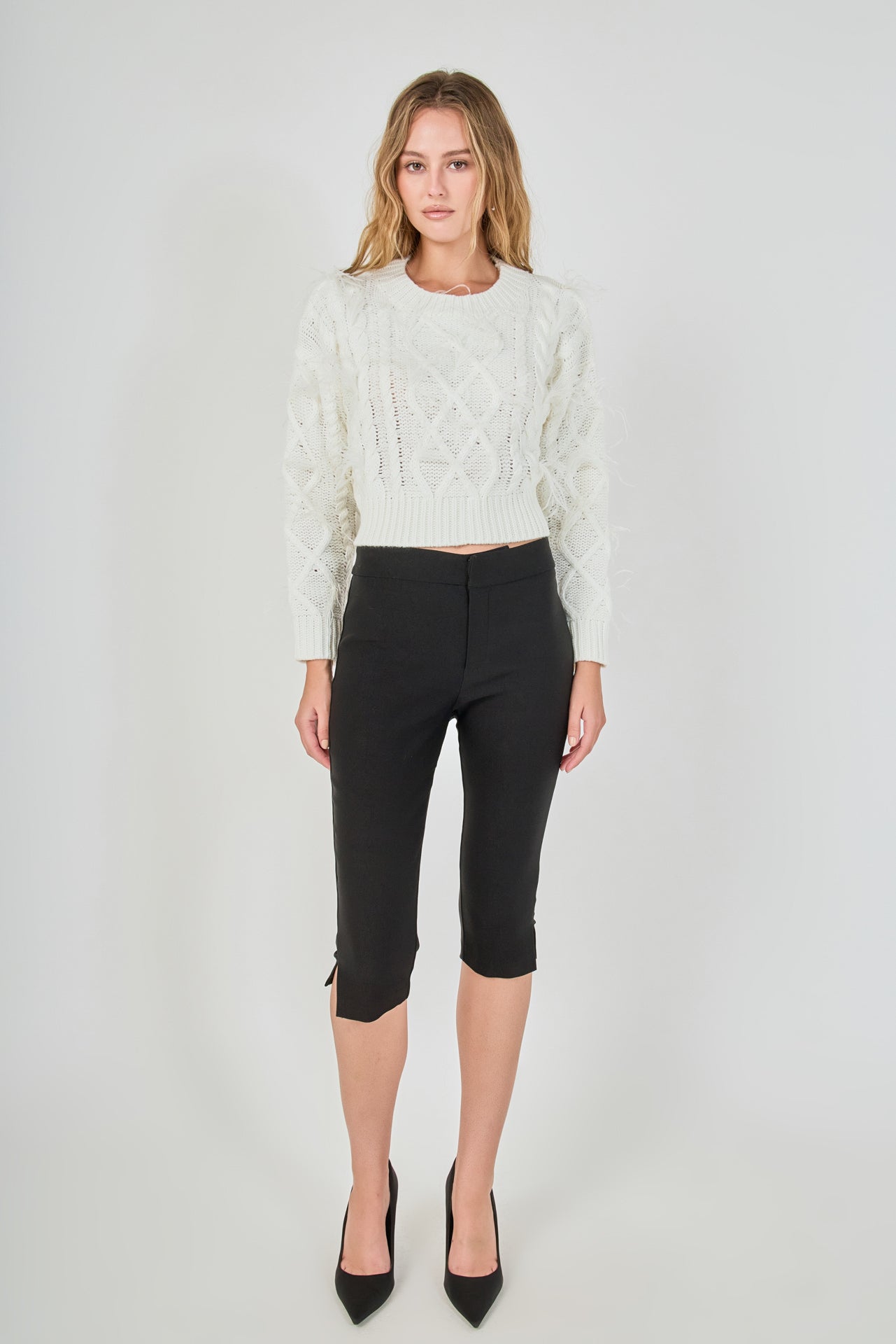 Full-length front view of a cream cropped cable-knit sweater with feather accents, styled with black fitted capri trousers.