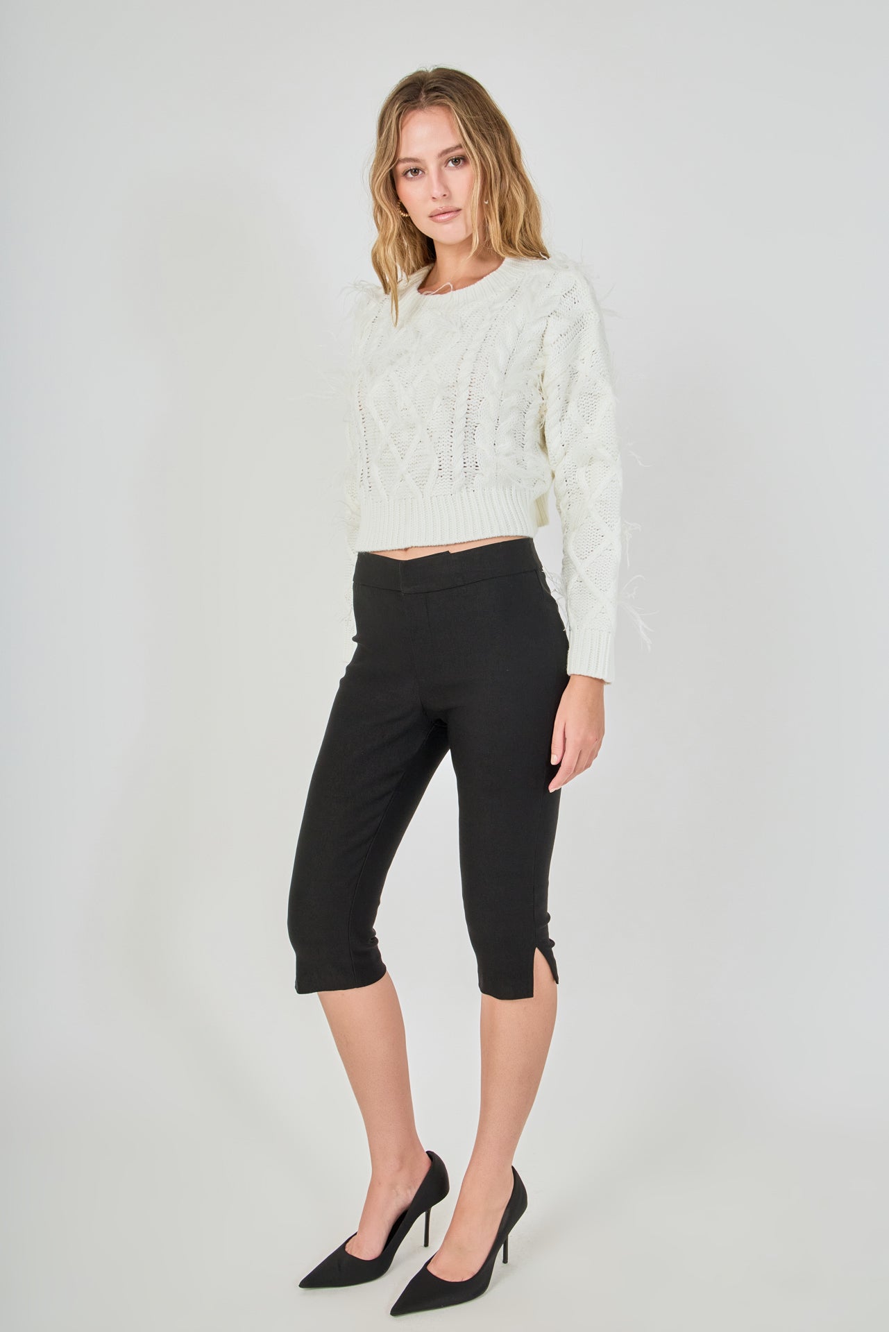 Side profile view of cream cropped cable-knit sweater displaying the long sleeve drape, wispy feather trim, and fitted waistline.