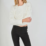 Face-forward posed view of a cream long-sleeve cable-knit cropped sweater featuring wispy feather accents woven throughout the cables.