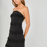 Three-quarter view of black strapless mini dress featuring horizontal floral appliqué trim, scalloped hem, and a fitted mini silhouette.