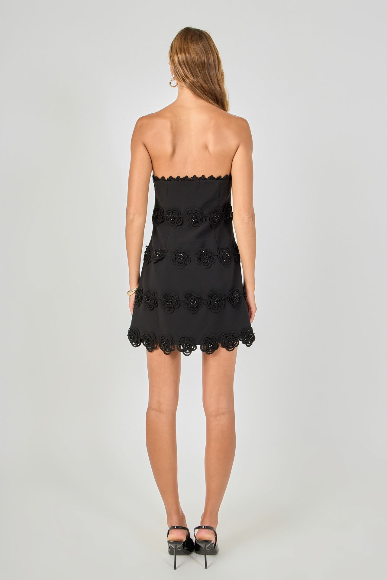 Back view of black strapless mini dress featuring horizontal floral appliqué trim and a straight hemline.