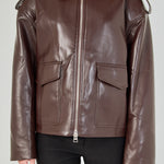 A woman wearing a dark brown faux-leather aviator-style jacket with a front zipper, shoulder epaulets, and flap pockets, paired with black pants, against a light gray studio background.