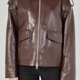 A woman wearing a dark brown faux-leather aviator-style jacket with a front zipper, shoulder epaulets, and flap pockets, paired with black pants, against a light gray studio background.