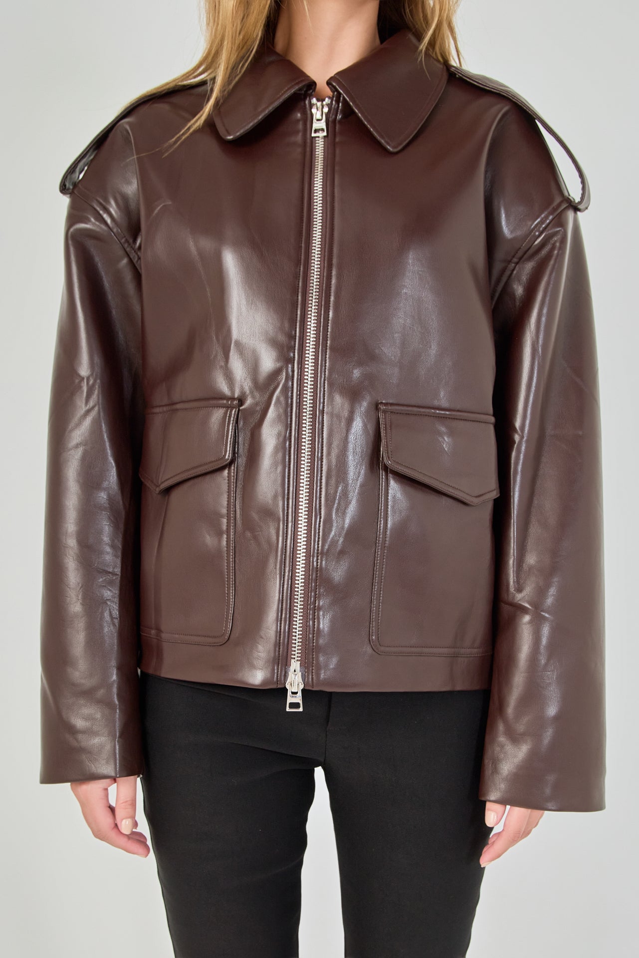 A woman wearing a dark brown faux-leather aviator-style jacket with a front zipper, shoulder epaulets, and flap pockets, paired with black pants, against a light gray studio background.