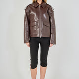 A woman modeling a brown faux-leather moto jacket with a front zipper, shoulder epaulets, and flap pockets, paired with black capri pants and black heels, against a light gray studio background.