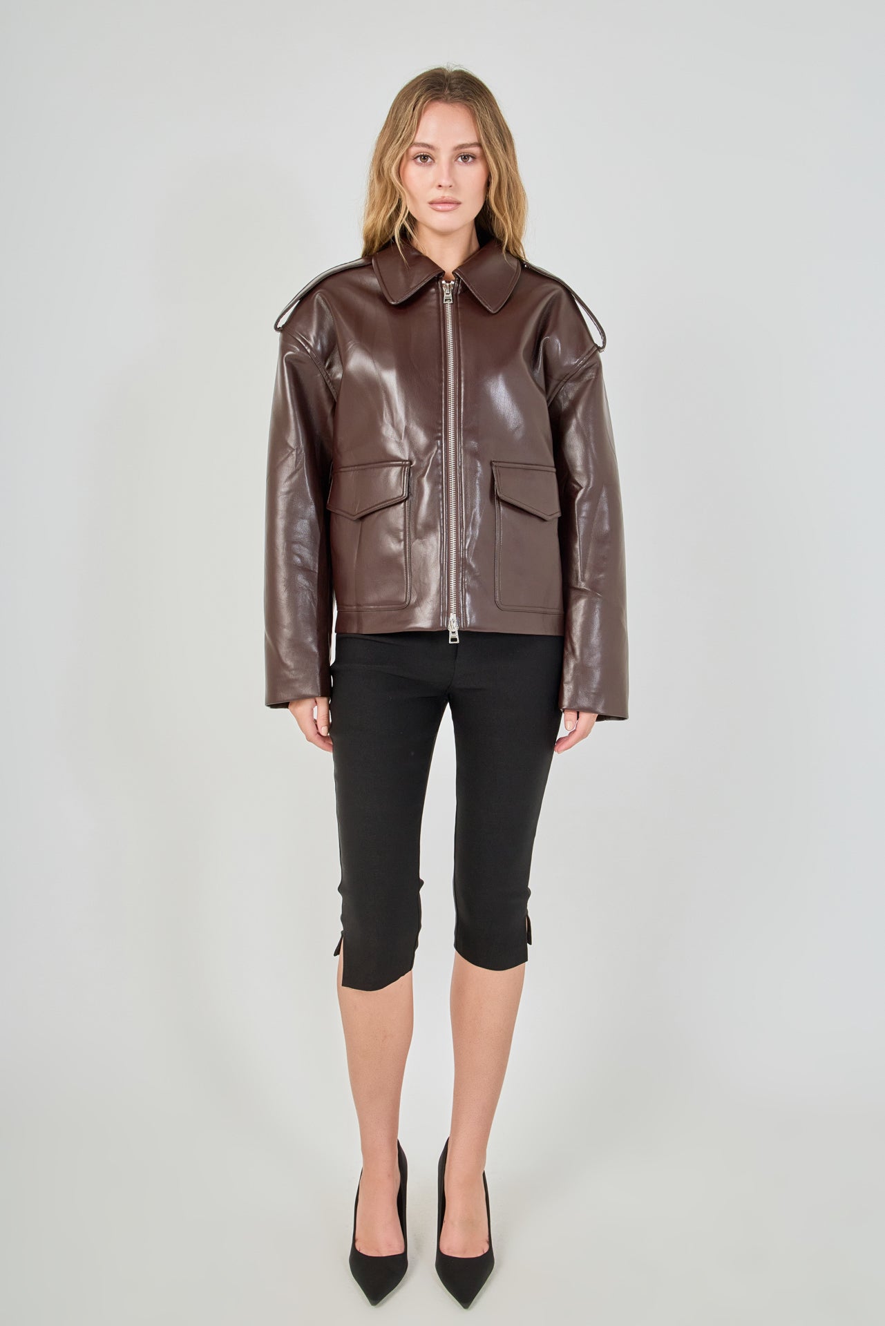 A woman modeling a brown faux-leather moto jacket with a front zipper, shoulder epaulets, and flap pockets, paired with black capri pants and black heels, against a light gray studio background.
