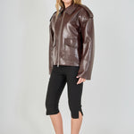 A woman modeling a brown faux-leather moto jacket with a collar and front zipper, paired with black cropped pants and black heels, against a light gray studio background.