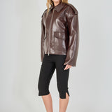 A woman modeling a brown faux-leather moto jacket with a collar and front zipper, paired with black cropped pants and black heels, against a light gray studio background.