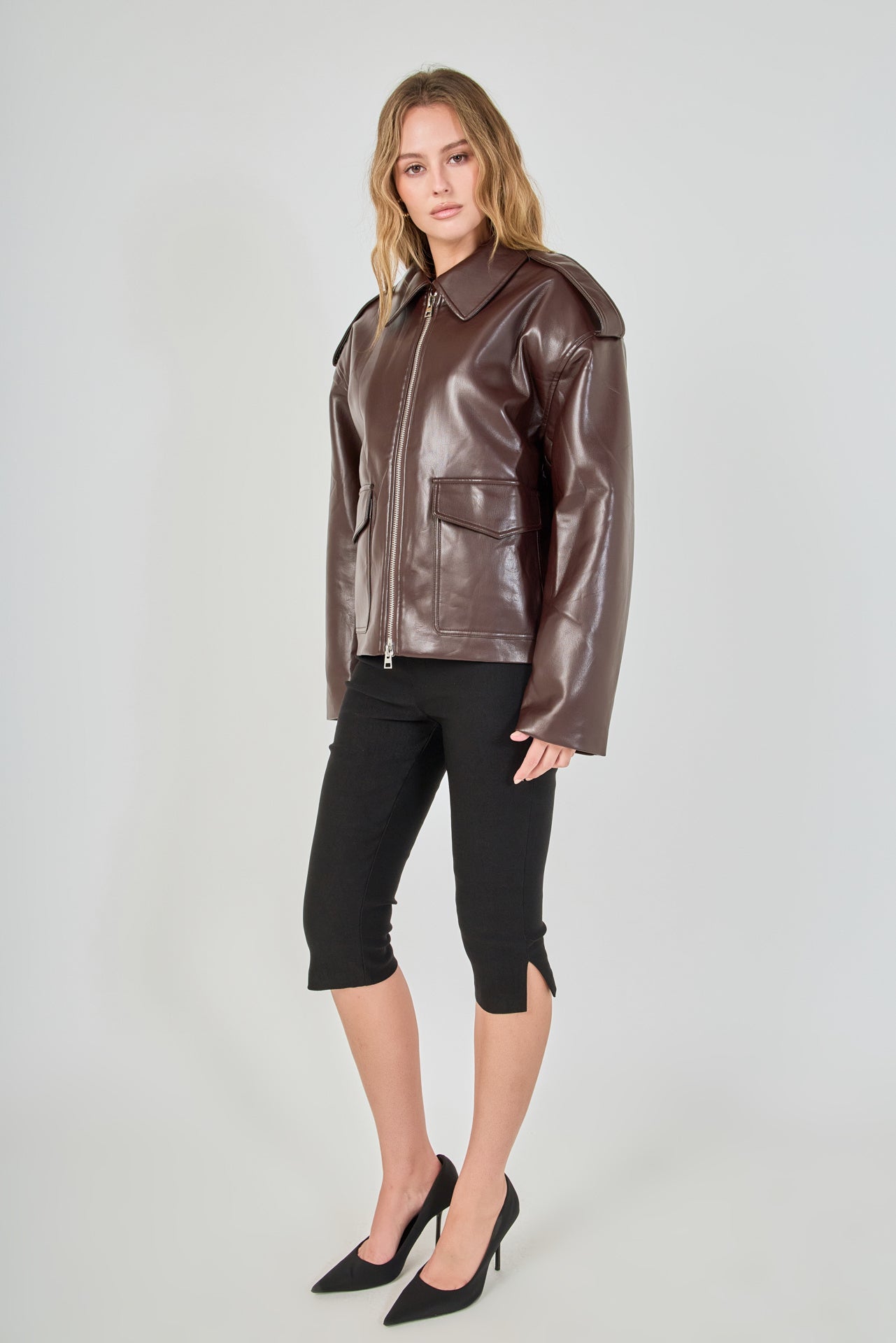 A woman modeling a brown faux-leather moto jacket with a collar and front zipper, paired with black cropped pants and black heels, against a light gray studio background.