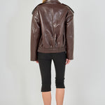 A woman viewed from the back wearing a brown faux-leather moto jacket, black cropped leggings, and black heels against a light gray studio background.