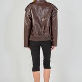 A woman viewed from the back wearing a brown faux-leather moto jacket, black cropped leggings, and black heels against a light gray studio background.