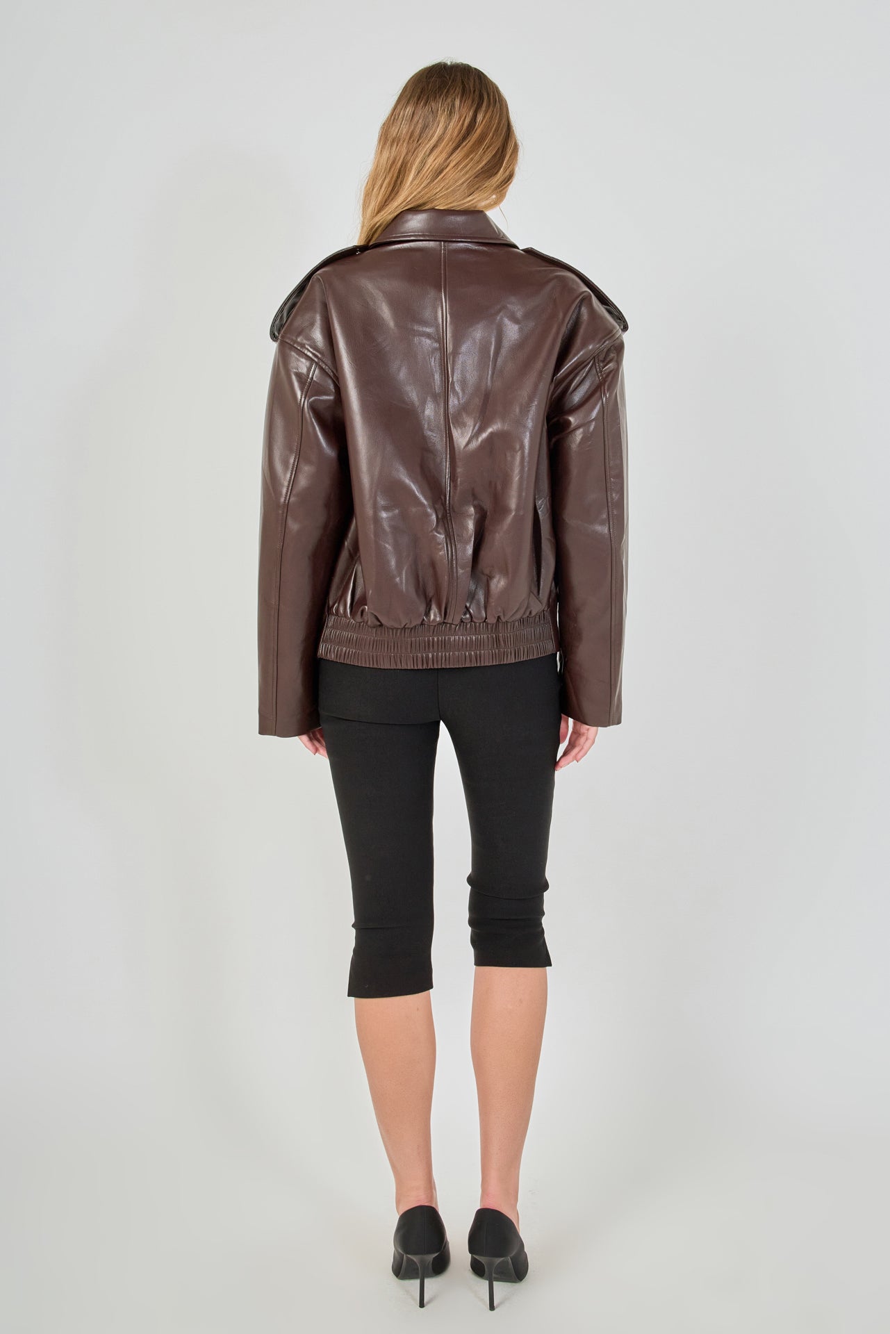 A woman viewed from the back wearing a brown faux-leather moto jacket, black cropped leggings, and black heels against a light gray studio background.