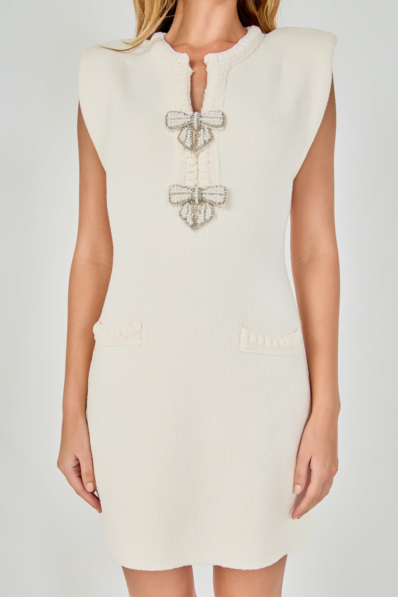 Upper body close-up view of a cream sleeveless knit mini dress showing the split neckline, cap shoulders, pearl bow embellishments, and faux hip pockets.