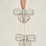 Ultra close-up detail view of cream knit fabric highlighting the front placket adorned with two ornate pearl-and-crystal bow brooches.